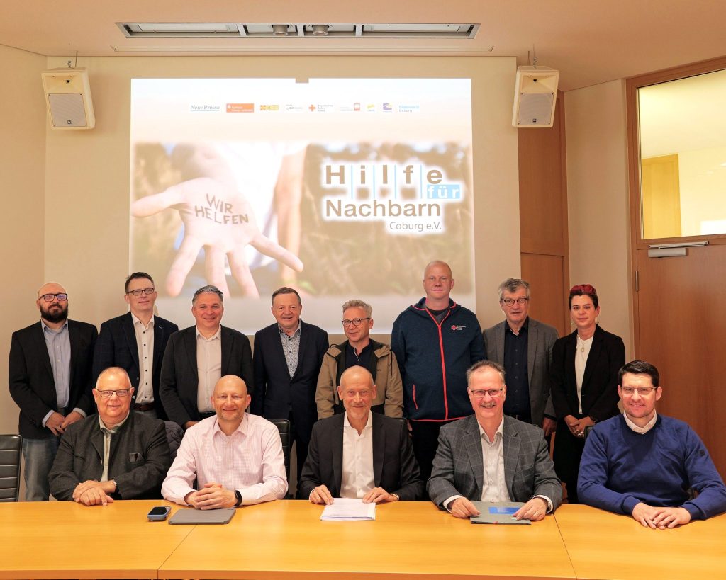 Group of men in suits posing for a photo in a conference room with a projected banner reading 'Hilfe für Nachbarn Coburg e.V.' in the background.