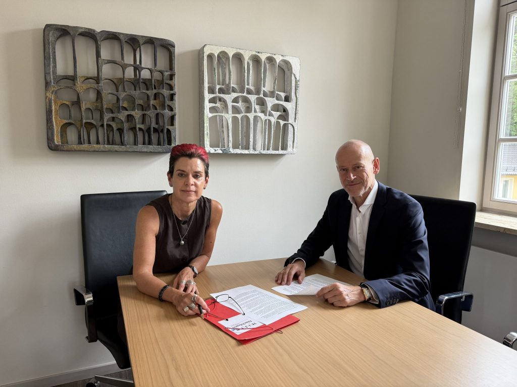 Two professionals sit at a light wood conference table, reviewing documents and red folders in a bright office.